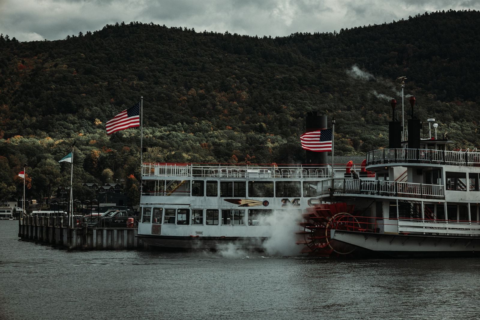 A paddlewheel steamboat flying American flags at a dock on Lake George, below wooded hills